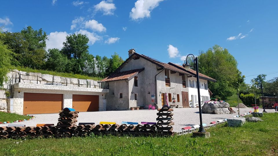 Cascina rurale ristrutturata con tetto in coppi e facciata chiara, affacciata su un ampio cortile in ghiaia con lampioni e piccoli elementi colorati lungo il bordo. A sinistra si vedono autorimesse con portoni in legno sotto un terrazzamento in pietra, circondati da prato e alberi.