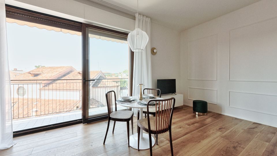 Soggiorno moderno e luminoso con pavimento in legno, pareti bianche a boiserie e zona pranzo con tavolo rotondo e sedie. Ampia portafinestra scorrevole con tende che apre su un balcone con ringhiera e vista sui tetti, con lampada a sospensione e piccolo mobile con TV.