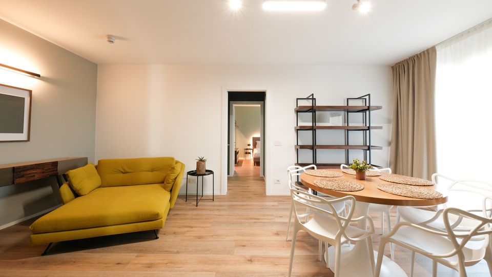 Soggiorno moderno e luminoso con pavimento in legno, divano chaise longue verde, tavolo da pranzo rotondo con sedie bianche e grande finestra con tende chiare. Sulla parete ci sono mensole in metallo e legno e una porta centrale che conduce ad altre stanze.