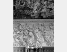 Parete interna decorata con un grande affresco in due registri, con scene figurative di stile classico e numerosi personaggi. In basso si vede un piccolo mobile con candele davanti alla parete, in un ambiente di tipo museale o storico.