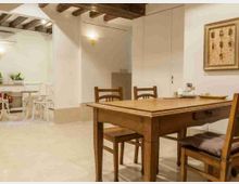 Interno di una sala da pranzo in stile rustico con tavolo e sedie in legno, soffitto con travi a vista e pareti chiare. Sullo sfondo si vede un piccolo angolo cucina con tavolo e sedie bianche e una parete con quadro incorniciato.