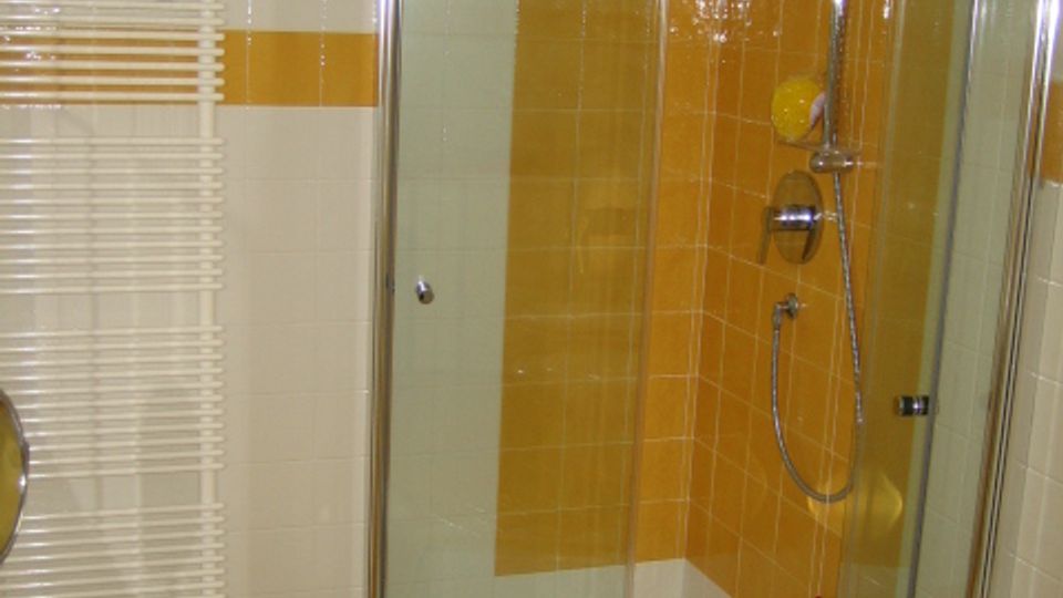 Bagno con box doccia ad angolo in vetro e profili metallici, piatto doccia bianco e soffione con doccetta. Pareti rivestite di piastrelle bianche con fasce arancioni e termosifone a scaletta sulla sinistra.