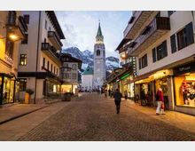 Strada pedonale lastricata in un centro alpino, fiancheggiata da edifici con balconi e negozi illuminati. In fondo si vede un’alta chiesa con campanile e guglie verdi, con montagne innevate sullo sfondo.