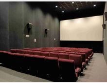 Interno di una sala cinematografica con file di poltrone rosse disposte a gradinata rivolte verso un grande schermo bianco. Pareti scure con altoparlanti laterali e soffitto a griglia con luci incassate.