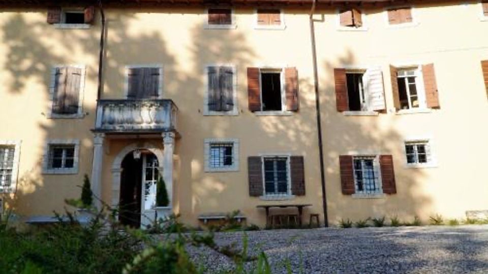Facciata di una casa rurale a due piani intonacata in giallo, con finestre con scuri in legno e un ingresso ad arco con piccolo balcone soprastante. Davanti si vede un cortile in ghiaia con alcune piante e ombre di alberi proiettate sul muro.