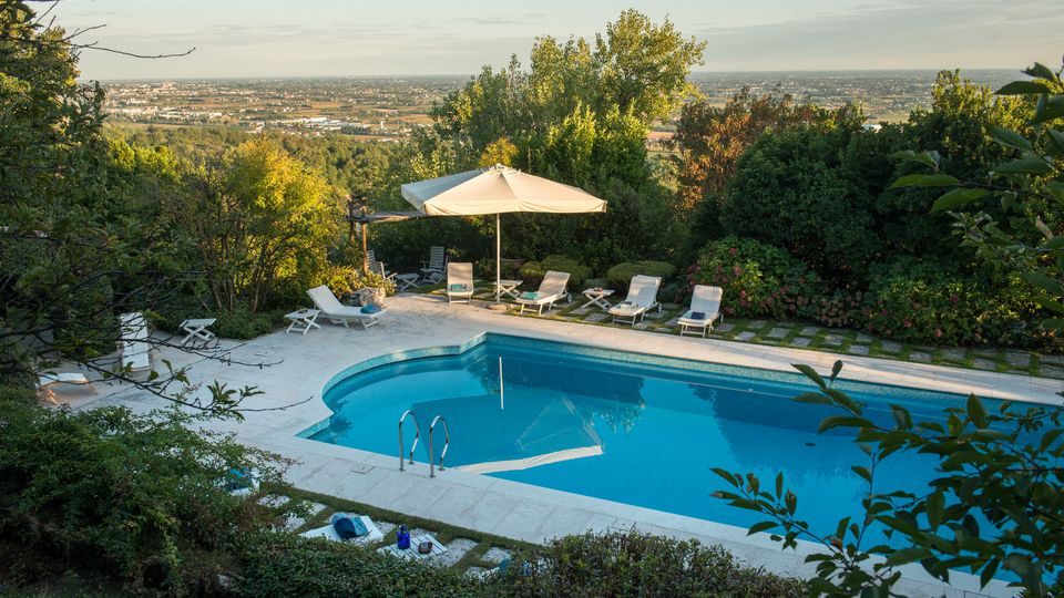 Piscina all’aperto con bordo in pietra chiara e acqua azzurra, circondata da siepi e alberi su un pendio. Intorno ci sono lettini e un grande ombrellone, con vista panoramica sulla pianura e su un’area urbana in lontananza.