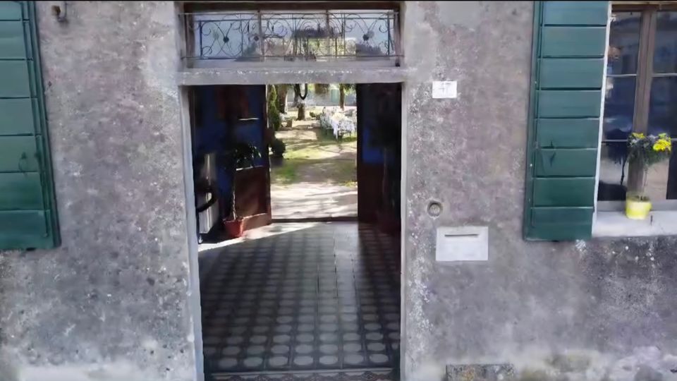 Ingresso ad arco di una casa con facciata in intonaco grigio e persiane verdi, con portone aperto su un corridoio pavimentato a piastrelle. Attraverso il passaggio si intravede un cortile interno con vialetto e vegetazione.