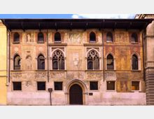 Facciata di un palazzo storico con intonaco decorato da affreschi, finestre ad arco e bifore con elementi gotici. Al centro si trova un grande portale ad arco, con file di finestre rettangolari al piano inferiore e una lunga gronda sporgente sotto il tetto.