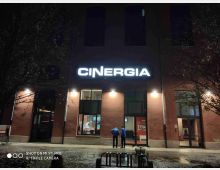 Facciata di un cinema urbano di notte con grande insegna luminosa “CINERGIA”, finestre alte e porte d’ingresso illuminate. Davanti all’edificio si vede il marciapiede bagnato e una piazza con panchina e alberi spogli ai lati.