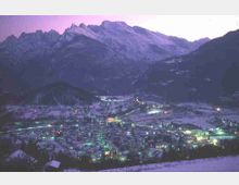 Veduta panoramica di una valle alpina al crepuscolo con un paese illuminato e campi innevati. Sullo sfondo si ergono alte montagne rocciose e pendii boscosi che incorniciano l’abitato.