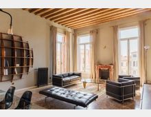 Soggiorno luminoso in stile contemporaneo con soffitto a travi in legno a vista, ampie finestre con tende chiare e pavimento in terrazzo. Arredi in pelle nera con tavolino in vetro su tappeto, libreria a parete e camino in marmo rosato vicino alle finestre.