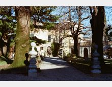 Viale di ghiaia in un giardino alberato che conduce a una villa chiara con portico ad archi e terrazza con balaustra. Ai lati del percorso si vedono grandi alberi e piedistalli in pietra con sfere decorative.