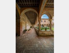Chiostro medievale in mattoni con portico ad arcate e colonne in pietra, pavimento in laterizio e soffitto a cassettoni decorato. Al centro si vede un giardino geometrico con siepi e vasi, circondato da logge e aperture ad arco verso il cortile interno.