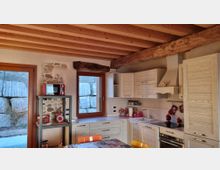 Cucina rustica con soffitto a travi di legno a vista e grande trave scura, mobili chiari a L con cappa e piano cottura. Due finestre e una porta finestra con cornici in legno affacciano su un muro in pietra; a sinistra una nicchia in pietra e una mensola metallica con piccoli elettrodomestici.