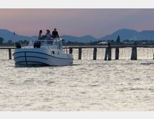 Piccola barca bianca su una laguna con acqua increspata, affiancata da una fila di pali di legno e reti che delimitano un’area. Sullo sfondo si vedono una costa bassa con vegetazione e profili di colline o montagne sotto un cielo al tramonto.