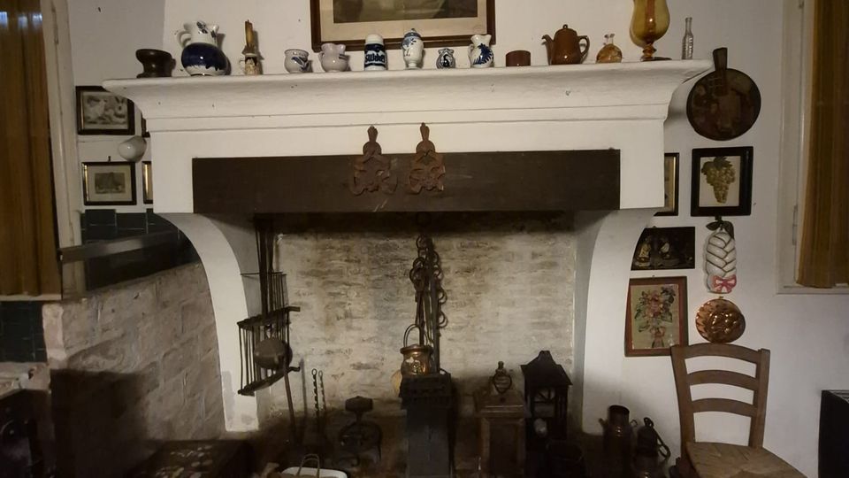 Interno rustico con grande camino in muratura bianca e focolare in pietra, con alari e attrezzi da camino. Sopra la mensola sono esposti piccoli vasi e oggetti decorativi, con un quadro incorniciato al centro e varie stampe e utensili appesi alle pareti laterali.
