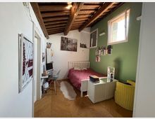 Camera da letto con soffitto in legno a travi a vista, parete verde e pavimento in parquet. Sono visibili un letto con copriletto rosso, una scrivania con sedia, mensole con libri e una finestra che illumina la stanza.