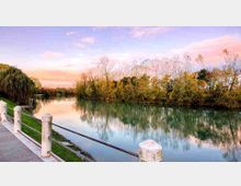 Passeggiata lungo la riva di un fiume calmo, con parapetto in metallo e colonnine in pietra sul lato sinistro. Sull’altra sponda si vede una fascia di alberi e vegetazione riflessa nell’acqua sotto un cielo al tramonto.