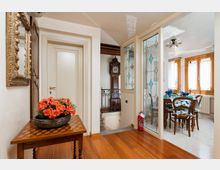 Ingresso interno di un’abitazione con pavimento in legno, tavolo consolle con composizione floreale e specchio dorato, porte bianche e una vetrata decorata che conduce alla zona pranzo. Sullo sfondo si vede una ringhiera in ferro e un orologio a pendolo, mentre la sala da pranzo è illuminata da grandi finestre con infissi in legno.