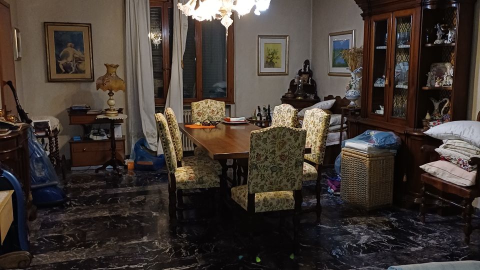 Ampia sala da pranzo in stile tradizionale con pavimento in marmo scuro, grande tavolo in legno con sedie imbottite e lampadario centrale. Alle pareti ci sono finestre con tende, quadri, un’alta credenza vetrata e vari mobili e oggetti domestici.