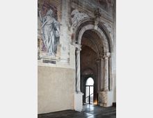 Interno di un edificio storico con grande arco in pietra sorretto da colonne marmoree e volta a botte, che conduce a un passaggio con porta ad arco vetrata. Le pareti sono decorate con affreschi e figure dipinte, tra cui una scena con un personaggio e iscrizioni.