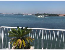 Terrazza sul mare con ringhiera bianca in primo piano e una palma in vaso. Sullo sfondo si vede un ampio tratto di laguna con un traghetto e altre imbarcazioni, oltre a una linea di costa con edifici e campanili all’orizzonte.