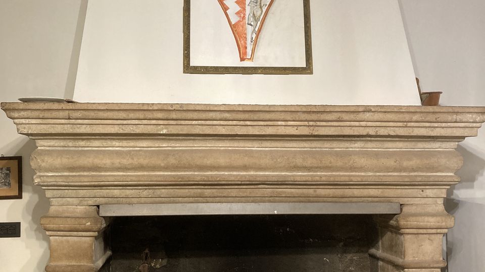 Grande camino in pietra con ampia cappa e focolare annerito, con alcuni utensili e recipienti in metallo e terracotta davanti. Sopra la mensola è appeso un pannello con uno stemma araldico su parete bianca.