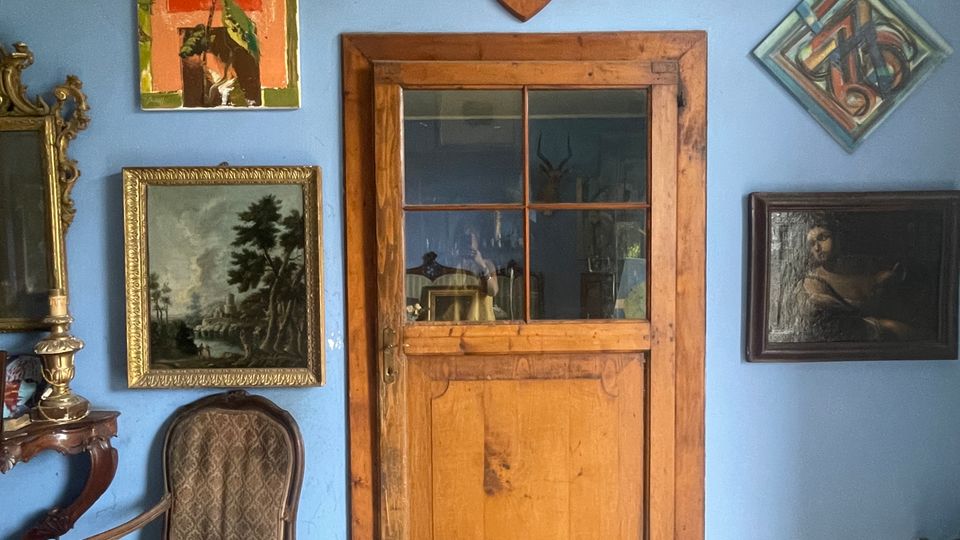 Interno di una stanza con parete azzurra e porta in legno con finestra a riquadri, circondata da quadri e cornici. Pavimento in piastrelle geometriche e arredi d’epoca, con una sedia imbottita e uno specchio su mobile lungo il lato sinistro.