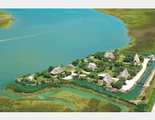 Veduta aerea di un piccolo villaggio su un’isoletta lungo un lago, con case dai tetti di paglia immerse nel verde e canali d’acqua che circondano la penisola. Si vedono pontili e barche vicino alla riva, con zone di palude e vegetazione acquatica tutt’intorno.