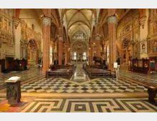 Interno di una grande chiesa con navata centrale, alte colonne e volte a crociera, con altare sul fondo. Pavimento a scacchiera e motivi geometrici in primo piano, file di banchi ai lati e pareti decorate con affreschi e cappelle.