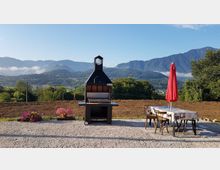 Terrazza in ghiaia con barbecue in muratura al centro e tavolo da esterno con sedie e ombrellone rosso sulla destra. Sullo sfondo si vede una vallata con alberi e campi e una catena montuosa sotto un cielo sereno.