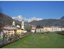 Veduta di una cittadina alpina con un ampio prato centrale affiancato da una fila di alberi potati e da edifici color pastello. Sullo sfondo si distinguono due campanili con cupole verdi e una catena di montagne rocciose innevate sotto un cielo sereno.