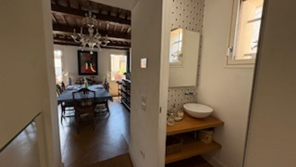 Interno di una casa con corridoio che conduce a una zona pranzo con tavolo e sedie, soffitto con travi a vista e lampadario. In primo piano c’è un piccolo bagno con lavabo su mensola in legno, parete a piastrelle decorate e una finestra.