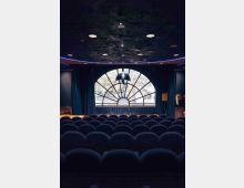 Interno di una piccola sala teatro o cinema con file di poltrone blu rivolte verso un grande finestrone semicircolare a raggiera. Soffitto scuro con luci incassate e pareti con tende blu che incorniciano la finestra.