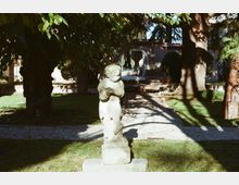 Giardino alberato con prato e vialetto in ghiaia, al centro una statua in pietra su piedistallo. Sullo sfondo si vede un edificio con arco d’ingresso e tetto in tegole, parzialmente ombreggiato dagli alberi.