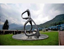 Grande scultura astratta in metallo su basamento circolare al centro di un prato, con edifici moderni sullo sfondo. Intorno si vedono colline boscose e montagne sotto un cielo nuvoloso.