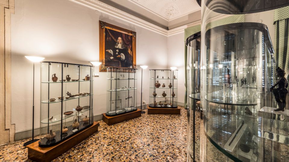 Sala museale con pavimento in terrazzo e soffitto decorato, con più vetrine in vetro e metallo che espongono piccoli reperti. Sulla parete di fondo è appeso un grande ritratto incorniciato e a destra si vede una vetrina cilindrica alta accanto a tende verdi.