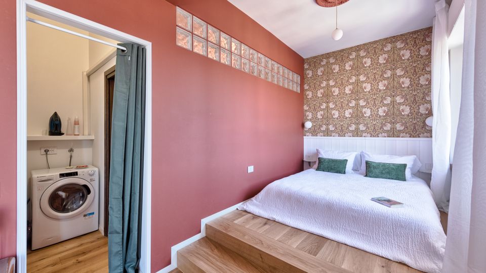 Camera da letto moderna con letto su pedana rialzata in legno, parete color terracotta con fascia di mattoni di vetro e testata con carta da parati floreale. A sinistra si apre una nicchia lavanderia con lavatrice e tenda, mentre sulla destra una finestra con tende bianche illumina l’ambiente.