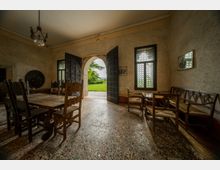 Interno di una sala rustica in stile storico con pavimento in graniglia, grande tavolo e sedie in legno, panca e piccola area con poltroncine. Una porta ad arco con grandi ante scure aperte conduce all’esterno su un prato verde, affiancata da finestre con vetri a piccoli riquadri.