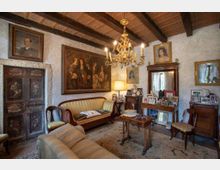 Salotto in stile tradizionale con soffitto a travi in legno e lampadario, pareti intonacate decorate da quadri e ritratti. La stanza contiene divani e sedie imbottite, un tavolino centrale, un mobile con specchio e una consolle con numerose cornici, oltre a una porta in legno scuro su una parete.