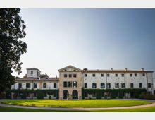 Facciata di una grande villa storica a più piani con tetto in tegole, finestre regolari e ingresso ad archi nella parte centrale. Davanti si estende un ampio prato verde curvo con vialetto e alberi ai lati, sotto un cielo sereno.