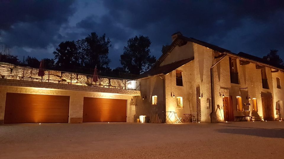 Edificio rurale in pietra illuminato di notte, con facciata a due piani, finestre e porte in legno e lampade a parete. A sinistra si vedono due ampie porte di garage sormontate da una terrazza con muretto in pietra e ombrelloni chiusi, sullo sfondo alberi e cielo nuvoloso.