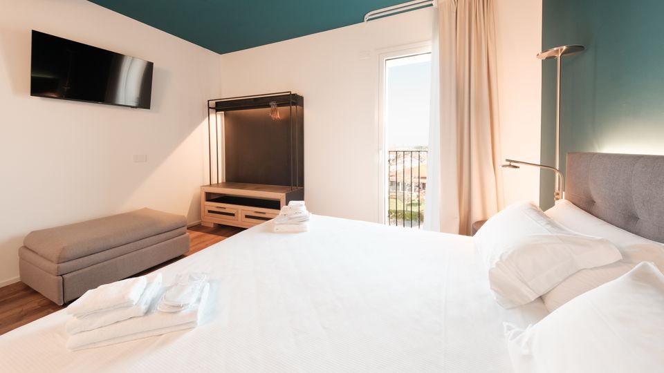 Camera da letto moderna con letto matrimoniale, pareti bianco e verde petrolio, TV a parete e mobile guardaroba aperto. Una portafinestra con tende chiare si apre su un piccolo balcone con ringhiera, illuminando la stanza con luce naturale.