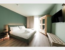 Camera da letto moderna con pareti verde salvia, letto matrimoniale con biancheria bianca e illuminazione a parete. Davanti a una finestra con tende chiare si trovano un armadio in legno con ante a vetro; sulla parete laterale sono presenti una TV a schermo piatto e una panca imbottita.