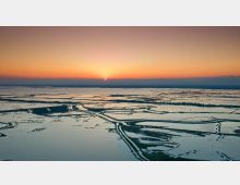 Veduta aerea di una vasta zona di laguna o palude costiera con canali e argini che formano geometrie irregolari sull’acqua. All’orizzonte si vede il sole al tramonto sotto un cielo sfumato tra arancione e blu.