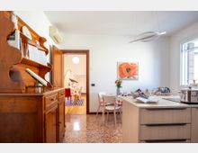 Cucina-sala da pranzo luminosa con pavimento in terrazzo, grande credenza in legno a sinistra e isola moderna con cassetti in primo piano. Tavolo con sedie sotto una finestra, lampada a sospensione circolare e quadro floreale sulla parete, con porta aperta verso un’altra stanza.