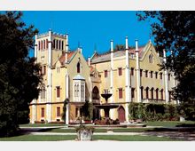 Edificio storico in stile neogotico con torre merlata, finestre ad arco e numerosi pinnacoli bianchi sul tetto. Davanti si vede un giardino con prato, vialetti e una fontana centrale, incorniciato da alberi.