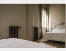 Camera da letto con pareti grigie, grande armadio a specchio e letto matrimoniale con testiera decorata. Una finestra lascia entrare luce naturale; si vedono anche un termosifone in ghisa e un piccolo comodino.