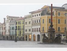 Piazza urbana sotto un forte temporale, con una fila di edifici storici color pastello a più piani e portici ad arcate al piano terra. In primo piano si vede una fontana monumentale con statue di leoni e un alto obelisco, mentre l’acqua piovana crea una nebbia sul selciato.