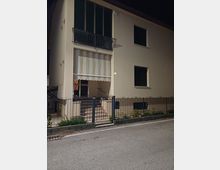 Facciata di una palazzina residenziale chiara vista di notte, con ingresso rialzato raggiungibile da una scalinata e una tenda a righe sopra la porta. Al piano superiore ci sono ampie finestre con balcone in ferro, e davanti all’edificio un cancello e una recinzione metallica lungo il marciapiede e la strada.
